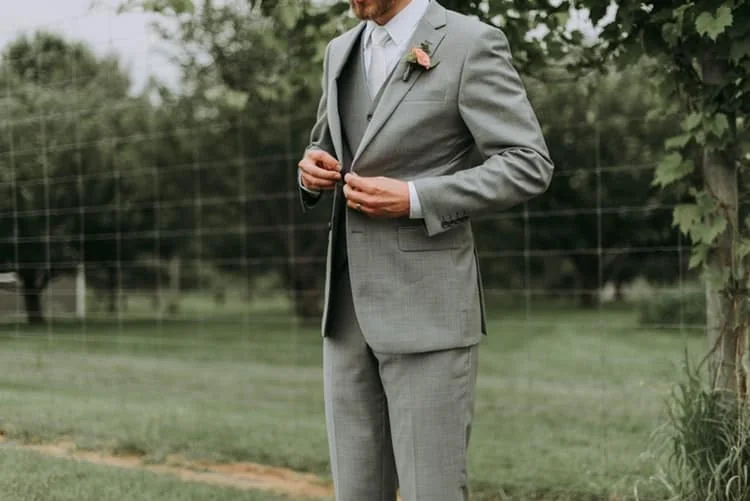 A member of the wedding party fastening a grey suit jacket with a buttonhole flower.