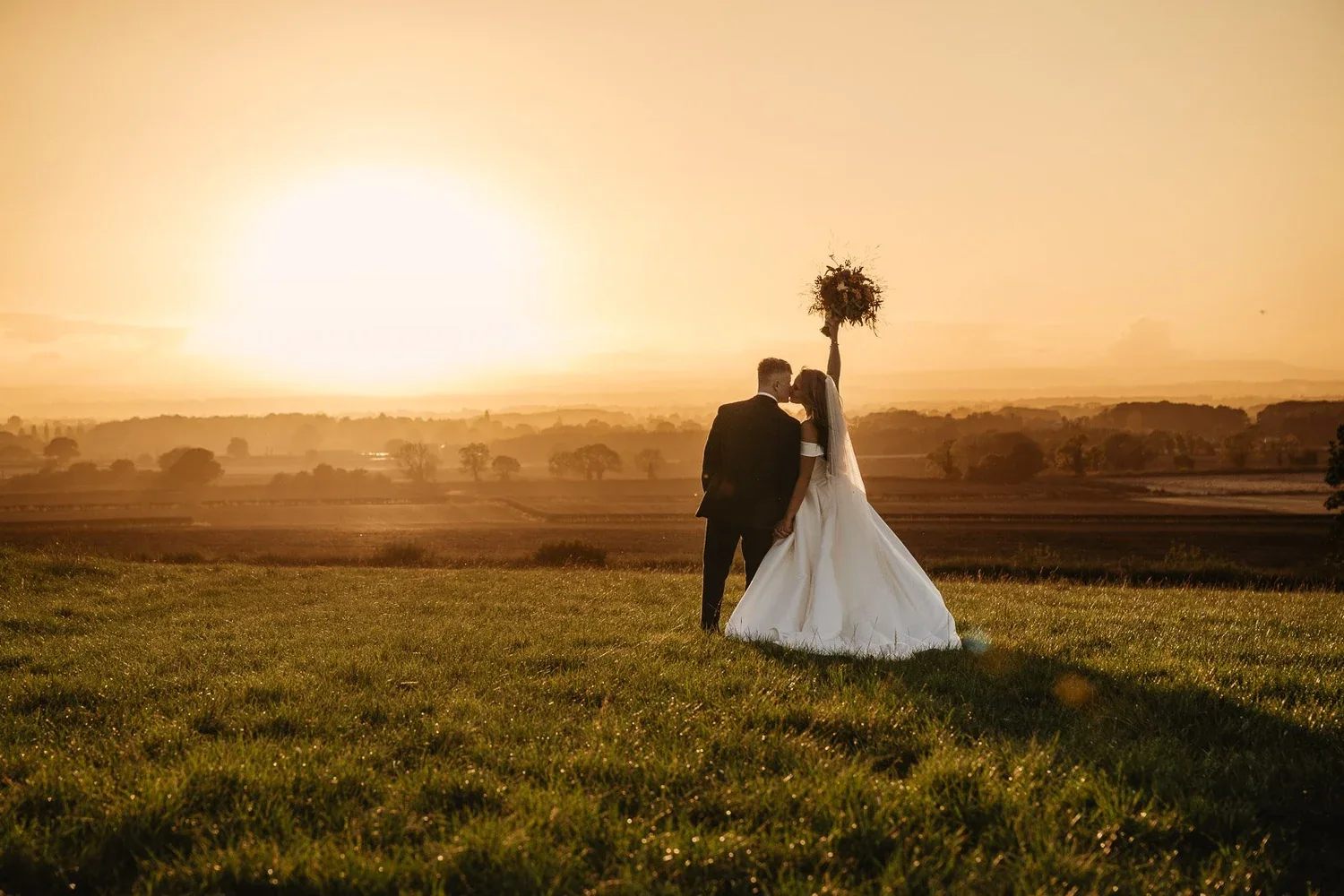 The bride raising her bouquet beside the groom against the setting sun.
