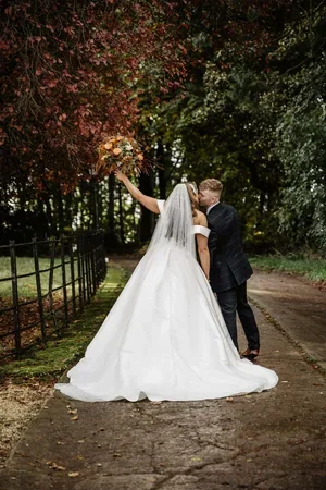The newlyweds kissing on a tree-lined path, with the bride's bouquet raised.