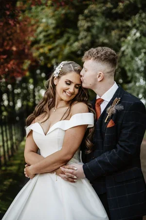 The groom kissing the bride's cheek as they stand beneath autumn leaves.