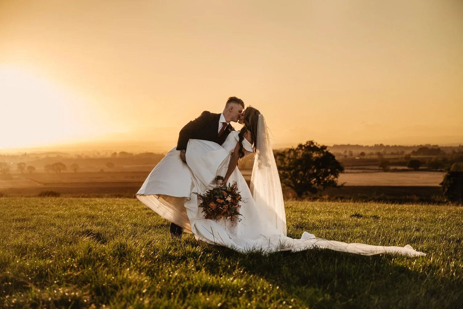 The groom dipping the bride for a kiss in the field at sunset.
