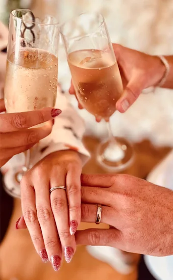 Wedding rings on the couple's hands beside raised glasses of sparkling wine.