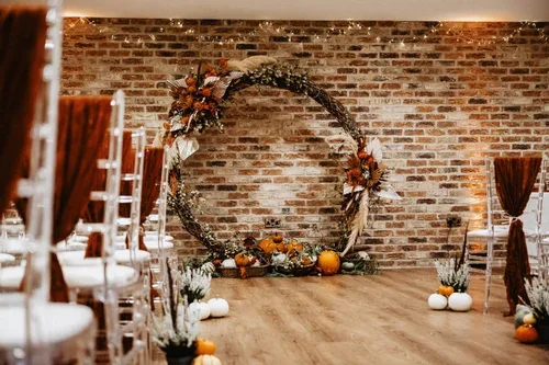 A circular floral ceremony arch with autumn leaves and pumpkins against a brick wall.