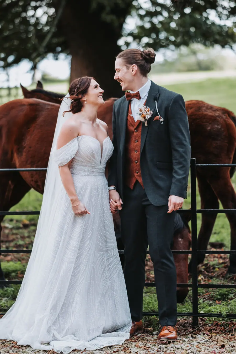 The couple holding hands beside two horses beneath the trees