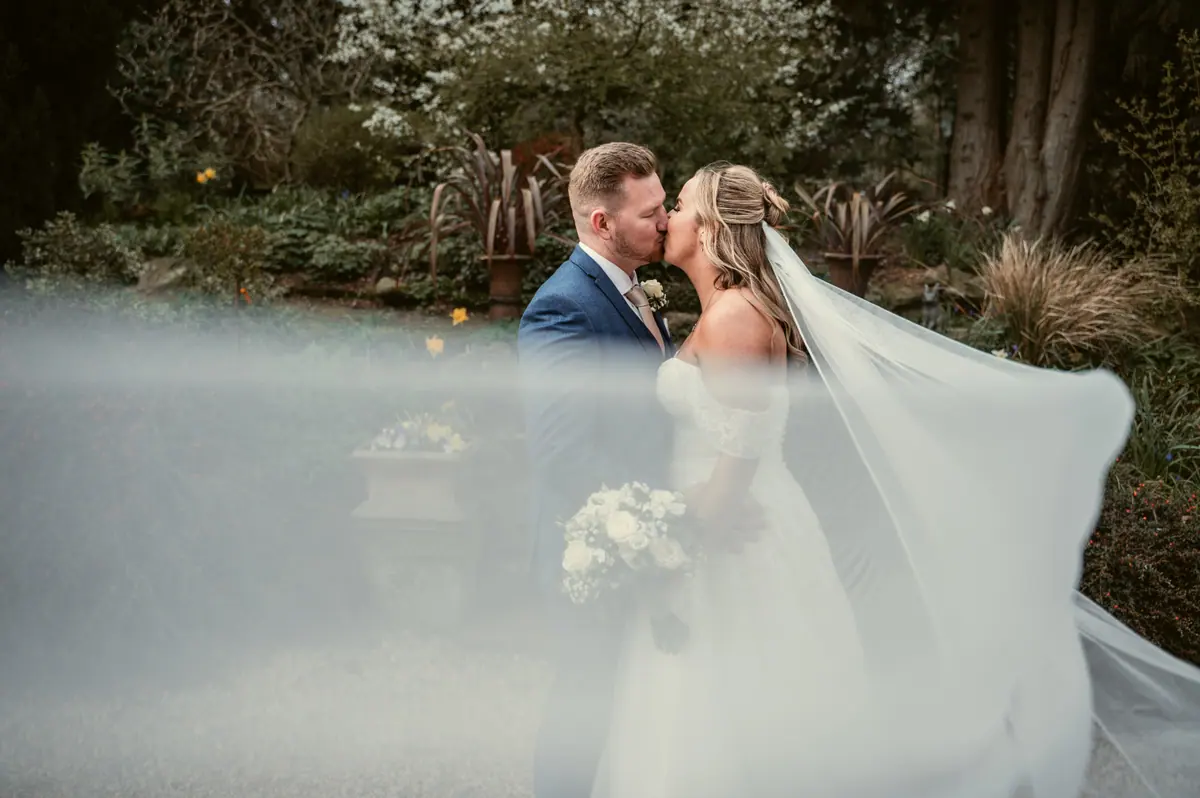 Hayley and Jayrrd kissing beneath her veil among garden trees
