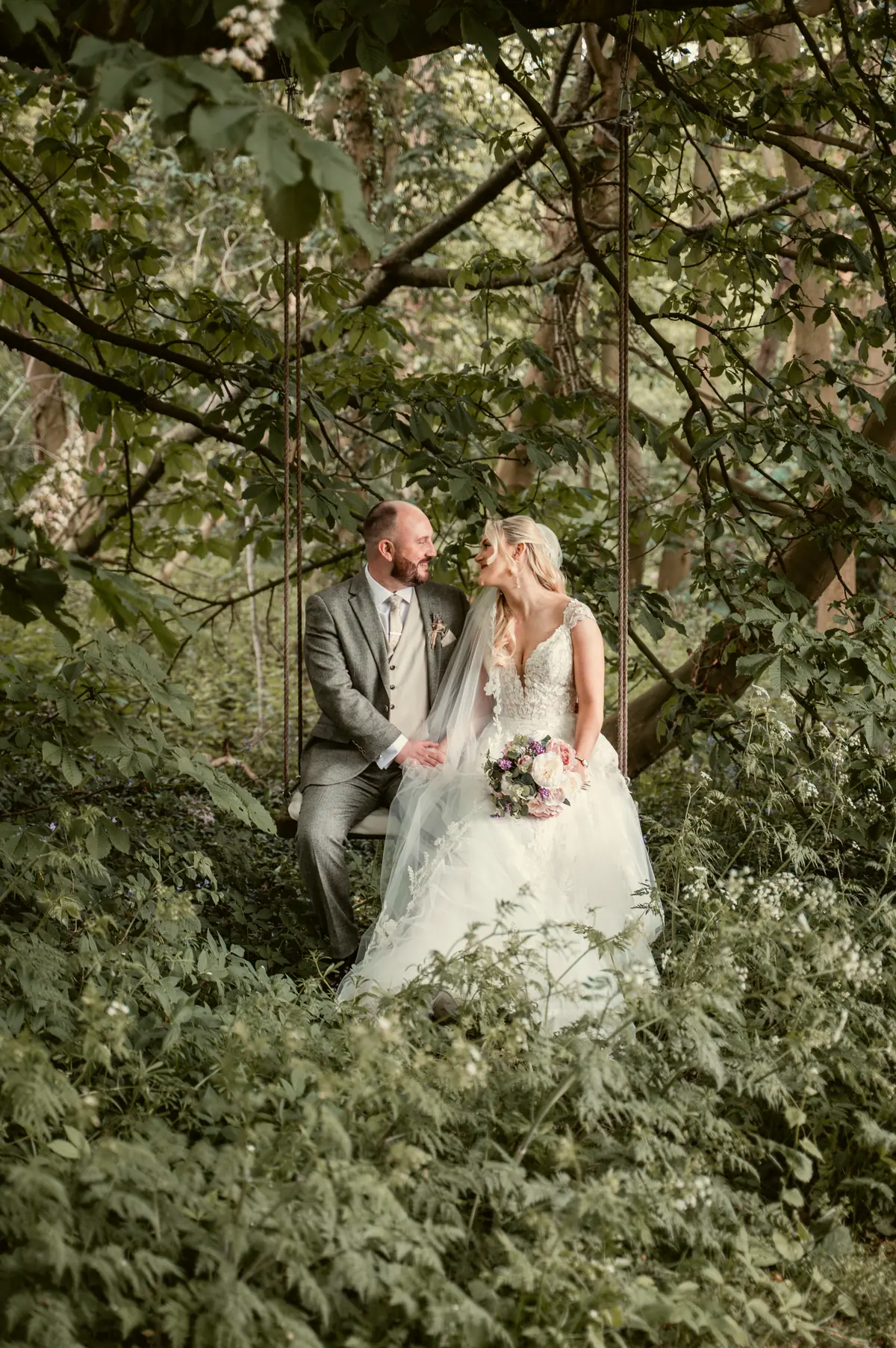 Katie and Chris embracing among leafy plants beneath woodland shade