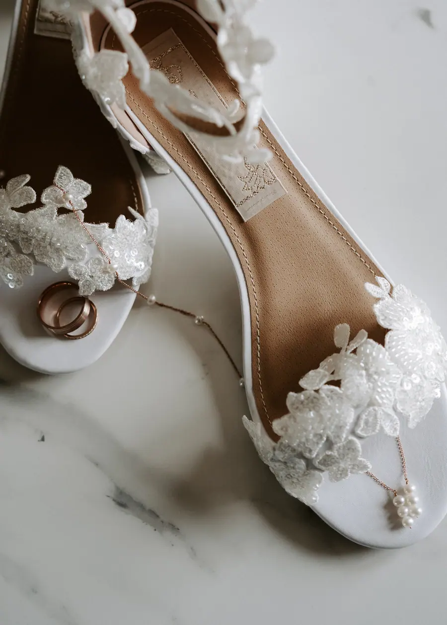 Lace wedding shoes, rings and pearl jewellery arranged on a pale surface