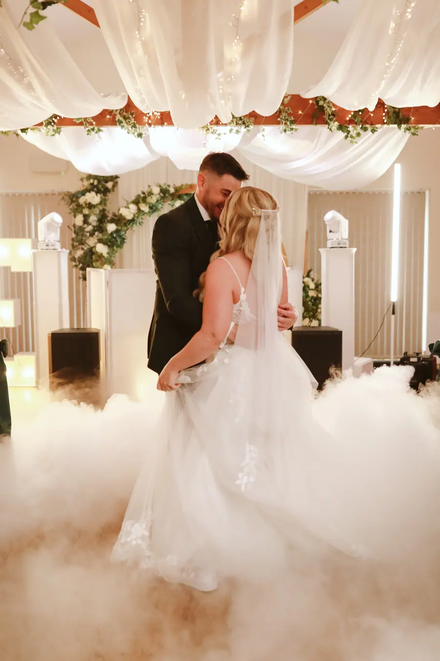 The couple dancing through low white fog beneath a circular floral arch