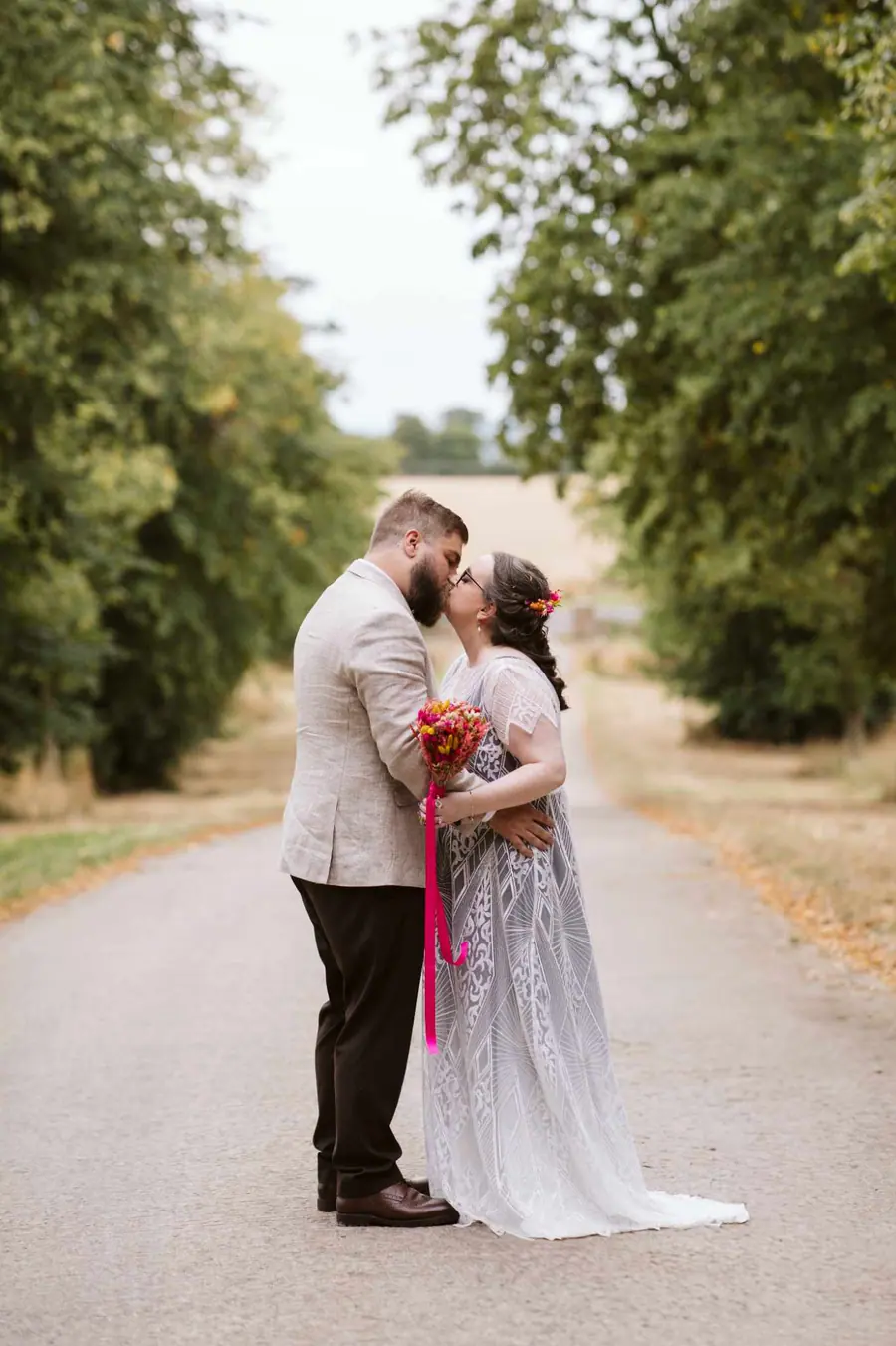 The couple kissing on the tree-lined lane with fields behind them