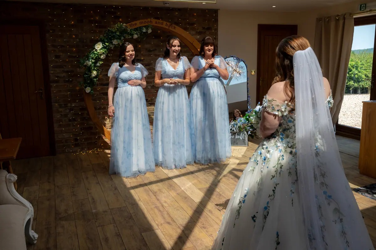Megan facing three women in pale blue dresses beside round mirrors