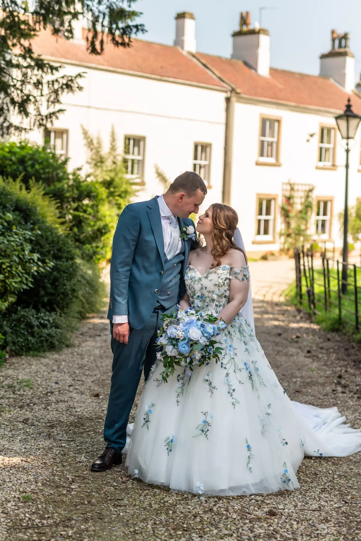 The couple kissing on gravel beside a pale house and garden