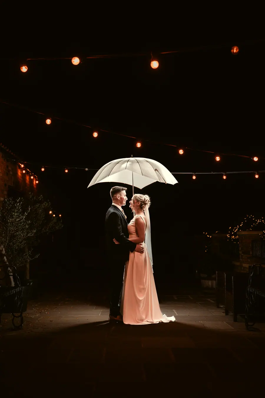 The couple standing beneath a white umbrella outside after dark