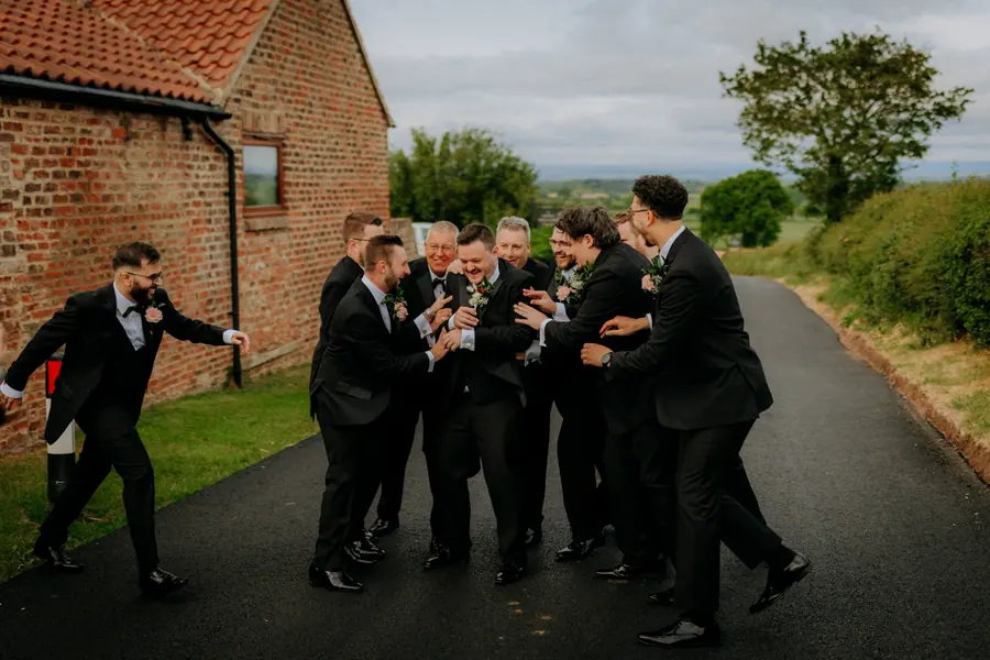 Men in black suits gathering playfully along the lane beside the barns