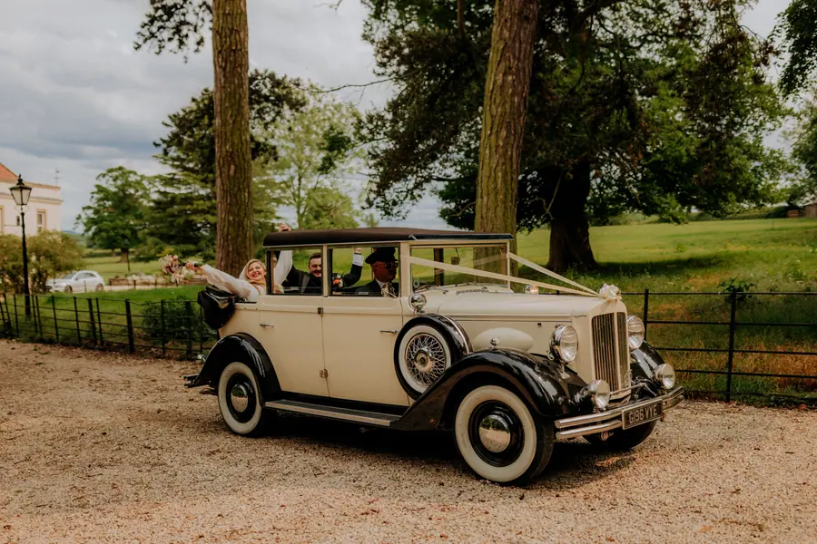 Cream vintage car parked beneath tall trees outside the venue