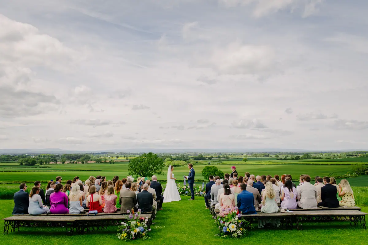 Guests seated on wooden benches for an outdoor ceremony overlooking fields