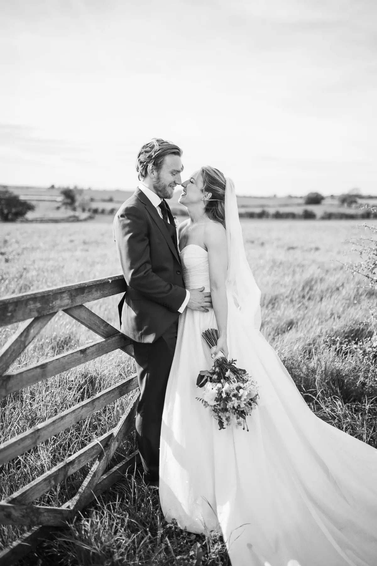 The couple standing at a field gate beneath a wide clouded sky