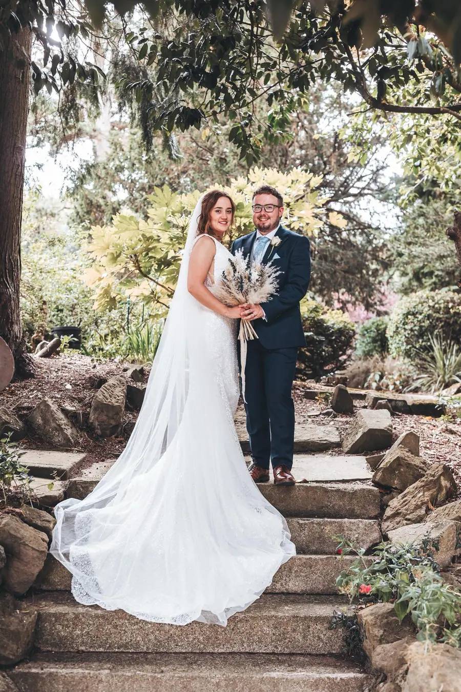 The couple standing together on stone steps beneath leafy branches