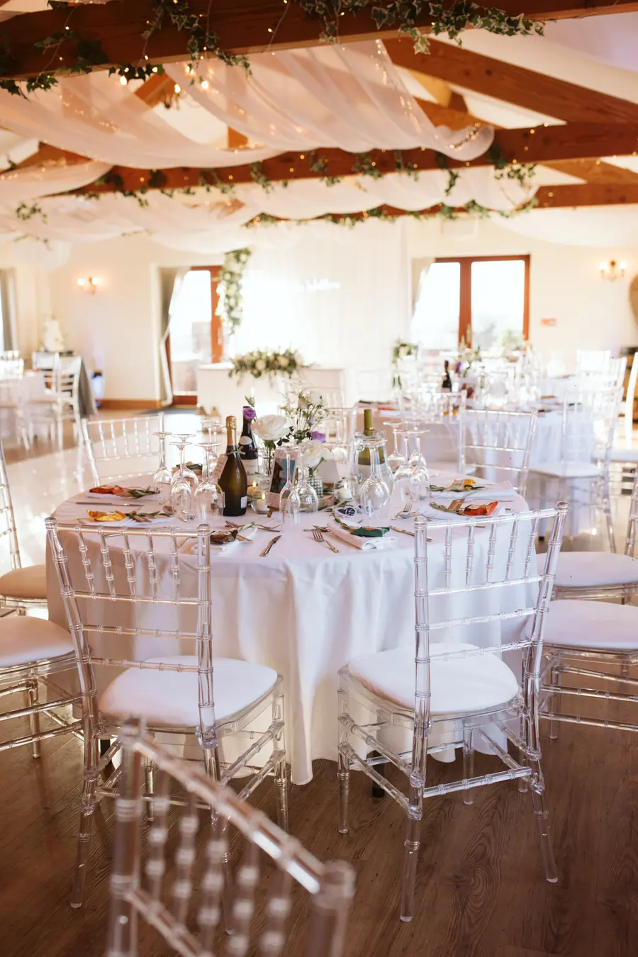 Round Hall tables dressed beneath white fabric, greenery and warm lights