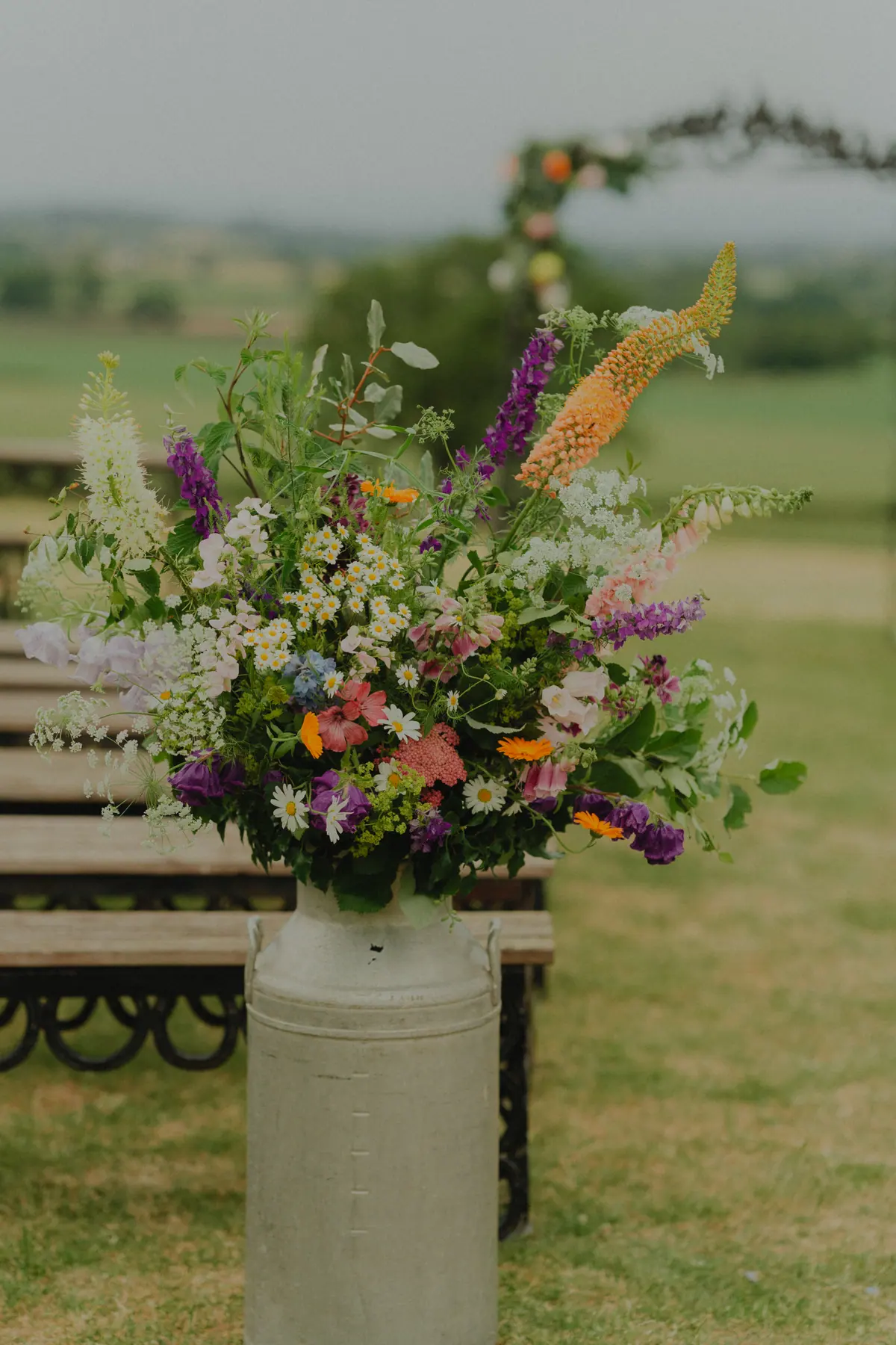 Colourful flowers arranged in a milk churn beside the outdoor ceremony benches