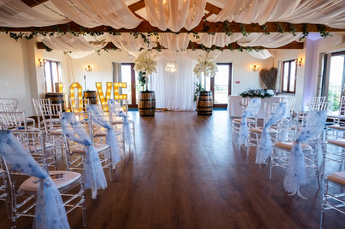 Clear ceremony chairs beneath soft ceiling drapes and greenery in the Hall