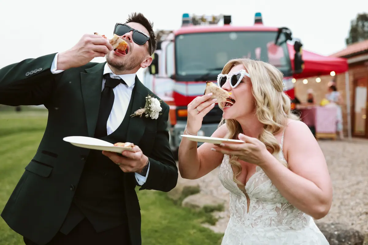 Newlyweds eating pizza in wedding sunglasses beside a red food truck at Thief Hall