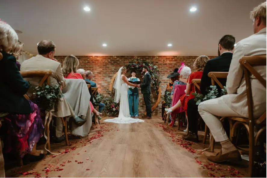 A couple holding hands with their celebrant at the end of the Small Hall aisle, surrounded by seated guests