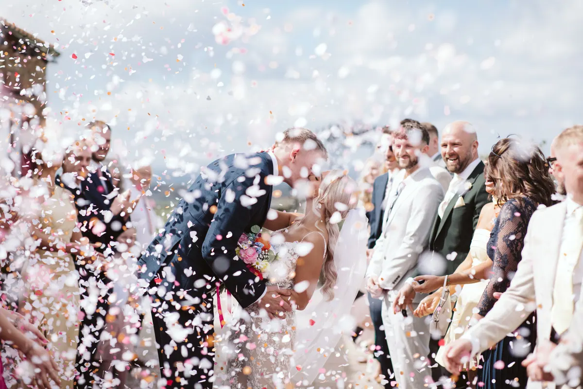Newlyweds kissing as guests throw confetti outside Thief Hall